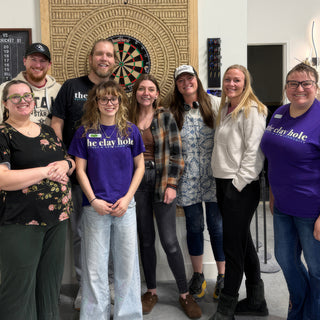 A group of potters standing shoulder to shoulder and smiling beside a dartboard, a few of them in purple t-shirts at The Clay Hole in Draper, Utah