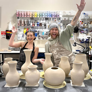 Two potters cheering with raised arms behind a table of unglazed vases in tall, round and bottle shapes at The Clay Hole in Draper, Utah