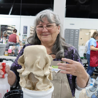 A smiling potter holding up a tan clay form with draping, tentacle-like folds above a white bucket at The Clay Hole in Draper, Utah