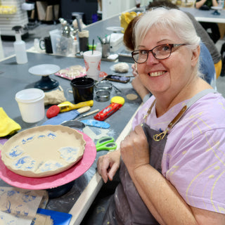 A smiling potter sits at a worktable beside a scalloped plate with blue patterned patches on a pink turntable at The Clay Hole in Draper, Utah