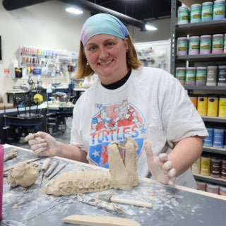 A potter in a pastel bandana smiling behind a long hand-built clay sculpture with rows of scales at The Clay Hole in Draper, Utah