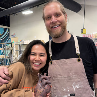 Two friends posing together, one holding a realistic sculpted heart in glossy dark brown, the other in an apron at The Clay Hole in Draper, Utah