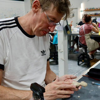 A potter looks down while shaping a small unfired clay figure in one hand at a work table at The Clay Hole in Draper, Utah