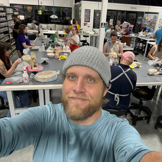 A potter in a gray beanie takes a selfie while a class works at long tables behind them at The Clay Hole in Draper, Utah