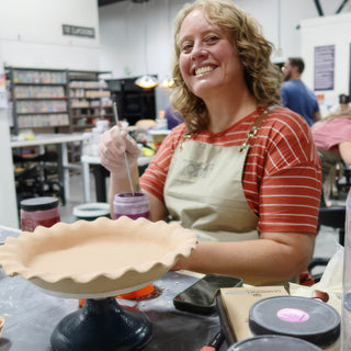 A smiling potter with a thin brush over a purple jar beside a shallow pale dish with a ruffled rim at The Clay Hole in Draper, Utah