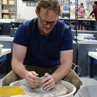 A potter in glasses and a navy t-shirt cups a gray clay form with both hands on the pottery wheel at The Clay Hole in Draper, Utah