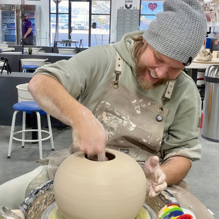 A laughing potter in a gray beanie reaching into the opening of a large round pale vessel on a wheel at The Clay Hole in Draper, Utah