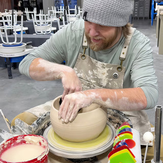 A potter reaching both hands into a wide, rounded clay pot on a pottery wheel with a bucket nearby at The Clay Hole in Draper, Utah