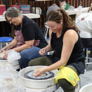 Two potters laughing at their wheels while the nearer one shapes a small clay bowl at The Clay Hole in Draper, Utah