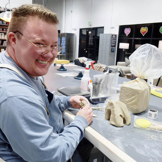 A smiling potter works on a small clay piece at a long worktable beside a plastic-wrapped block of clay at The Clay Hole in Draper, Utah