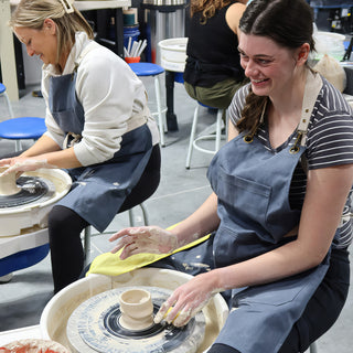 Two potters in aprons working at pottery wheels, the nearer one shaping a small clay cup with wet hands at The Clay Hole in Draper, Utah