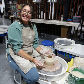 A smiling potter in a tan apron at a pottery wheel shaping a small tan clay vase with a tool at The Clay Hole in Draper, Utah