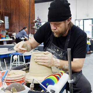 A potter in a black beanie and apron shaping the wall of a wide clay form on a pottery wheel at The Clay Hole in Draper, Utah