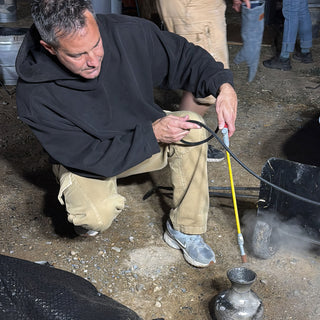 A person in a black hoodie kneels holding a yellow wand and hose beside a small shiny silver vase at The Clay Hole in Draper, Utah