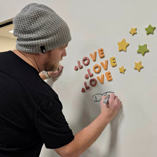 A person in a gray beanie signing a white wall with a marker beside clay LOVE letters and stars at The Clay Hole in Draper, Utah