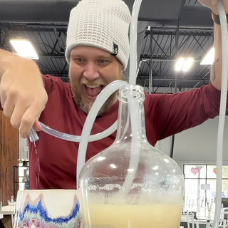 A person in a white knit beanie siphoning cloudy yellow liquid from a large glass jug through clear tubing at The Clay Hole in Draper, Utah