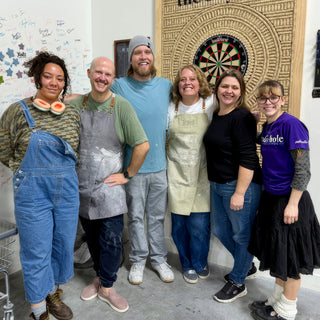 Six potters standing close together and smiling in front of a dartboard and a wall of handwritten signatures at The Clay Hole in Draper, Utah