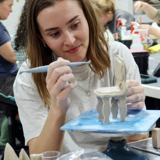 A potter carving fine detail into a small clay chair with curved legs using a long blue tool at The Clay Hole in Draper, Utah