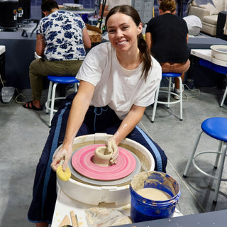A smiling potter holds a yellow sponge and shapes a small clay form on a pink wheel head at The Clay Hole in Draper, Utah