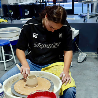 A potter in a black shirt shapes a small clay cup on a wheel, a yellow towel across their lap at The Clay Hole in Draper, Utah