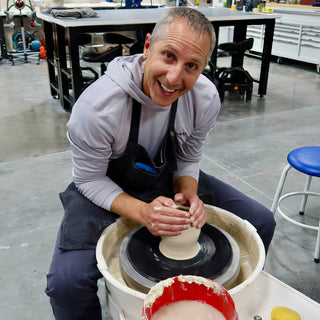 A smiling potter shapes a small pale clay cylinder on the pottery wheel inside a white splash pan at The Clay Hole in Draper, Utah