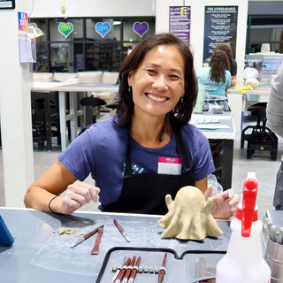 A smiling potter sits with a small pale clay ghost figure beside a tool roll and a spray bottle at The Clay Hole in Draper, Utah