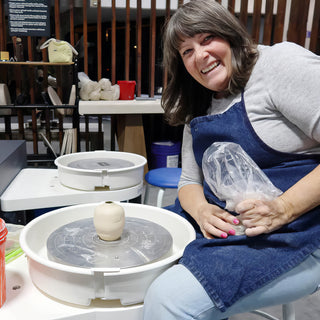 A smiling potter holds plastic-wrapped clay in their lap beside a wheel with a small cream vase on it at The Clay Hole in Draper, Utah