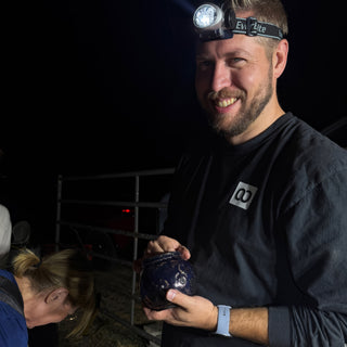 A smiling person in a headlamp holds a small round pot with a dark blue glossy finish at The Clay Hole in Draper, Utah
