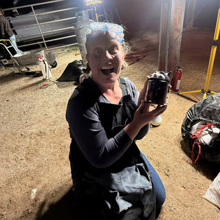 A potter kneels on dirt with goggles on their head and holds up a small dark glossy lidded jar at The Clay Hole in Draper, Utah