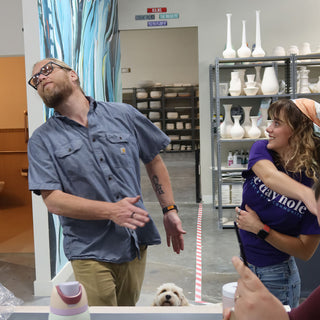 A small fluffy dog peers up from the bottom of the frame as two people move around the studio at The Clay Hole in Draper, Utah