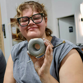 A smiling potter in thick-framed glasses holds up a small gray clay donut shape and a fine brush at The Clay Hole in Draper, Utah