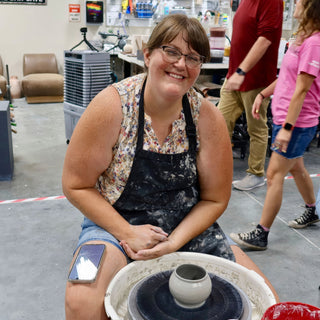 A smiling potter in a dark apron sits with folded hands at a wheel carrying a small round gray pot at The Clay Hole in Draper, Utah