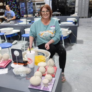 A smiling potter sits at a pottery wheel behind small pale lidded clay jars on pink and purple boards at The Clay Hole in Draper, Utah
