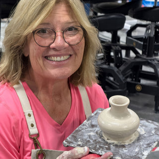 A smiling potter in a pink shirt holds a small pale gray vase with a narrow neck on a board at The Clay Hole in Draper, Utah
