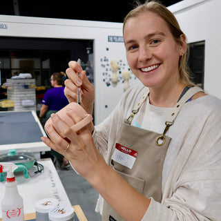 A smiling potter in a tan apron holds a small peach clay piece with a curved handle and a dropper at The Clay Hole in Draper, Utah