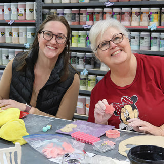 Two smiling potters at a table, one holding up a small pink clay cutout beside cutters and rolled clay at The Clay Hole in Draper, Utah
