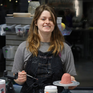 A smiling potter in a black apron holding a thin tool beside a small pink dome on a turntable at The Clay Hole in Draper, Utah