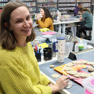 A smiling potter in a yellow green sweater works beside a board of small round clay discs with pressed patterns at The Clay Hole in Draper, Utah