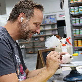 A smiling potter wearing a white earbud painting a small rounded clay piece with a thin brush on a turntable at The Clay Hole in Draper, Utah