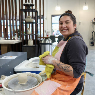 A potter in a colorful apron wiping hands on a yellow cloth by a small tan piece on a wheel at The Clay Hole in Draper, Utah