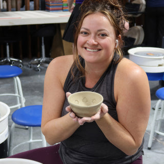 A smiling potter in a gray tank top holding a small unglazed bowl with small pressed marks inside it at The Clay Hole in Draper, Utah