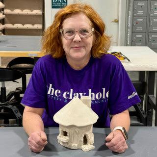 A potter in a purple shirt sits behind a small unglazed clay house with a chimney and arched door at The Clay Hole in Draper, Utah
