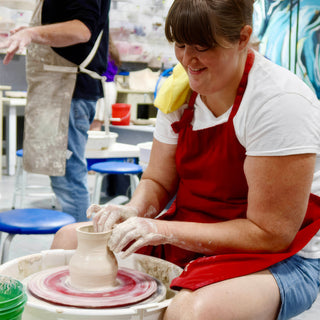 A potter in a red apron shaping a small rounded vase on a wheel with a pink swirled wheel head at The Clay Hole in Draper, Utah