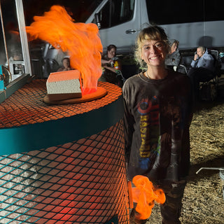 A smiling person in a graphic shirt stands beside a teal mesh kiln with flames rising from the top at The Clay Hole in Draper, Utah