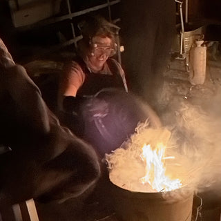 A smiling potter in safety goggles stands behind a metal barrel with flames and thick white smoke rising from it at The Clay Hole in Draper, Utah