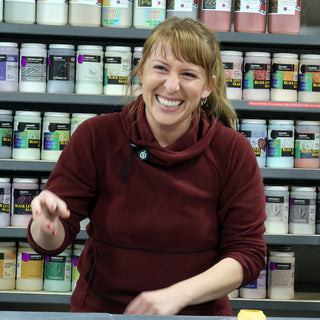 A laughing potter in a maroon pullover gestures in front of shelves of labeled glaze jars at The Clay Hole in Draper, Utah