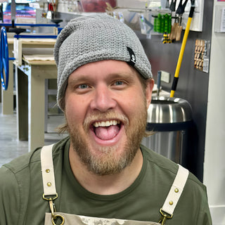 A potter in a gray knit beanie and olive apron grins wide with a wall of hanging tools behind at The Clay Hole in Draper, Utah