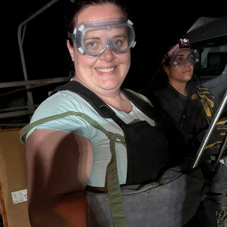 A smiling potter in clear safety goggles and a dark apron stands in front of another potter holding tongs at The Clay Hole in Draper, Utah