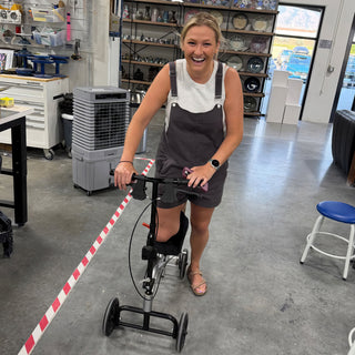A smiling person in gray overalls rests one leg on a knee scooter by a red and white taped line at The Clay Hole in Draper, Utah