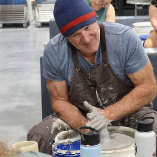 A potter in a blue beanie smiling at the camera with wet hands on clay on a pottery wheel at The Clay Hole in Draper, Utah